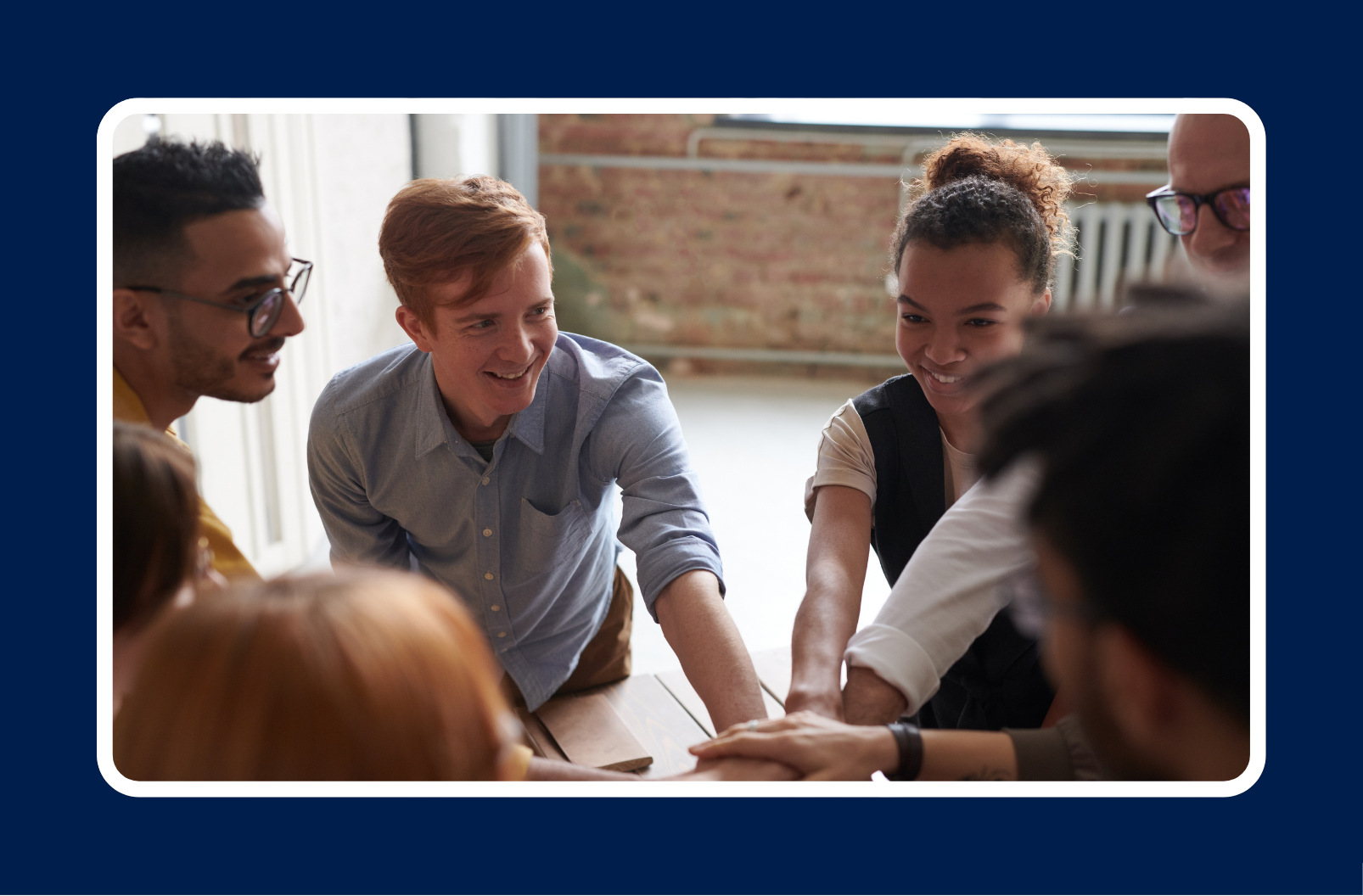 A group of young professionals smiling and stacking hands in a gesture of teamwork and unity, seated around a table in a casual office setting with natural light.