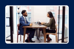 "Two professionals sitting across from each other at a table in a modern office, engaged in a job interview or business meeting. Both have laptops open, and one is holding a resume or document. The setting includes large windows and a clean, minimalist design.
