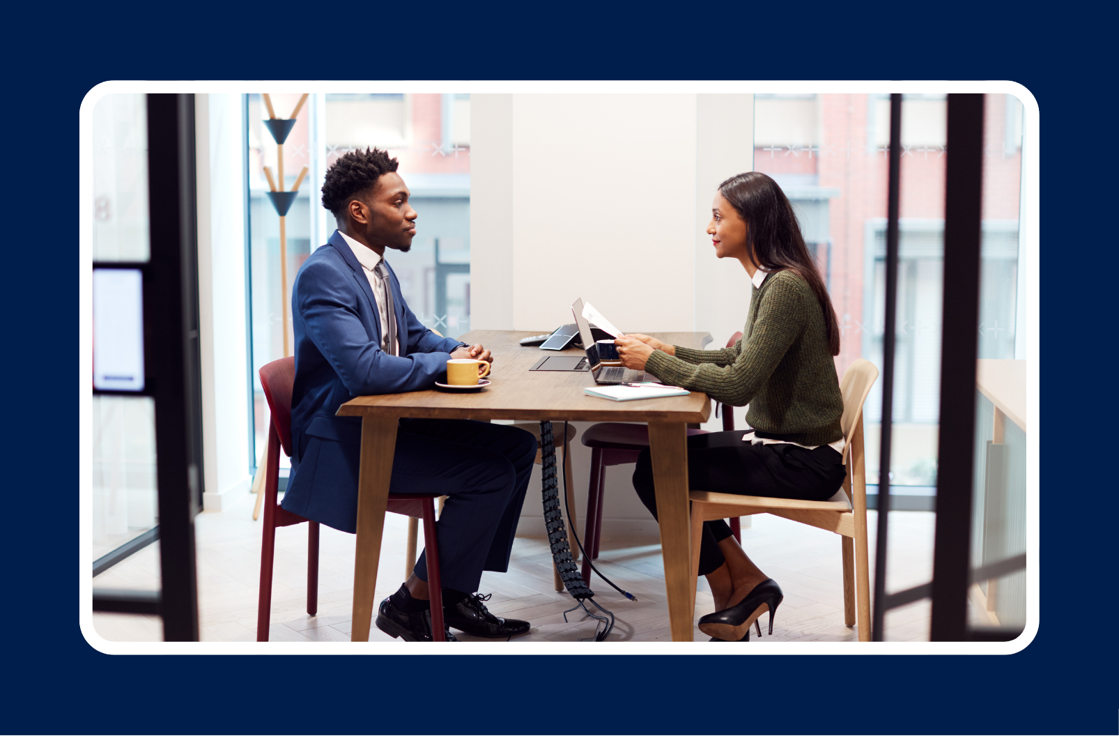 "Two professionals sitting across from each other at a table in a modern office, engaged in a job interview or business meeting. Both have laptops open, and one is holding a resume or document. The setting includes large windows and a clean, minimalist design.