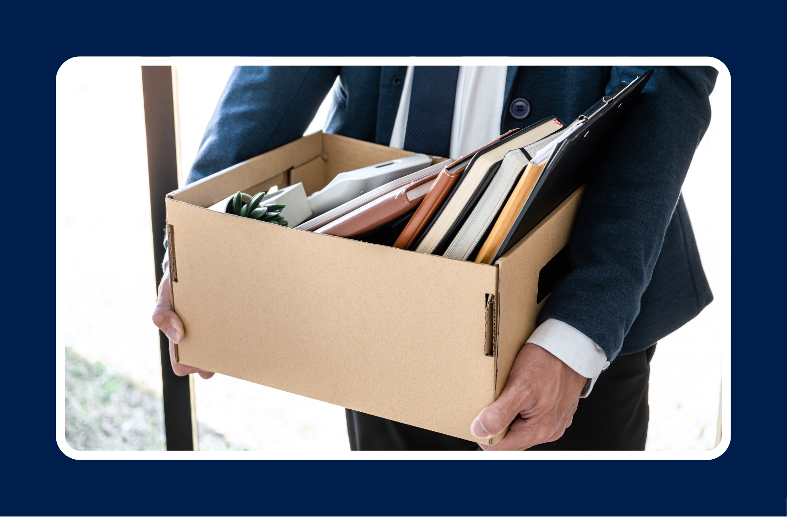 A person in professional attire holding a cardboard box filled with office supplies and personal items, suggesting a job change or layoff.