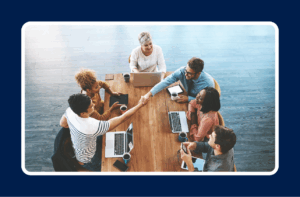 A diverse group of professionals sitting around a table with laptops, shaking hands and collaborating on a project, representing IT onboarding best practices for team integration and new hire success.