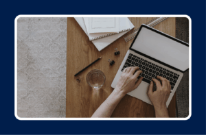 Person working on a laptop at a wooden desk with notebooks, pencil, and glass of water, representing remote work in full-time vs contract IT employment.