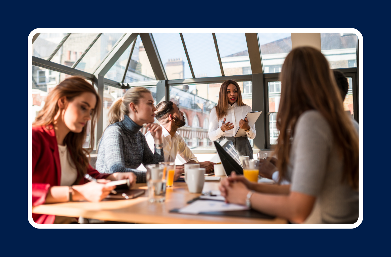 A group of professionals participates in a team meeting inside a modern glass-walled office as a woman leads the discussion with a tablet in hand, illustrating effective cybersecurity training for employees and collaborative workplace learning.