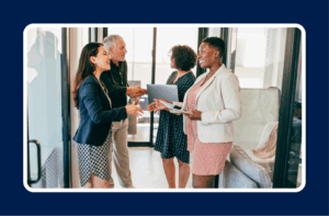 A group of professionally dressed colleagues stand in a modern office hallway, smiling and shaking hands during a networking-style conversation. Two people hold laptops and tablets as they discuss work. The scene reflects collaboration and highlights common IT hiring bottlenecks such as communication gaps, slow processes, and misalignment between teams.
